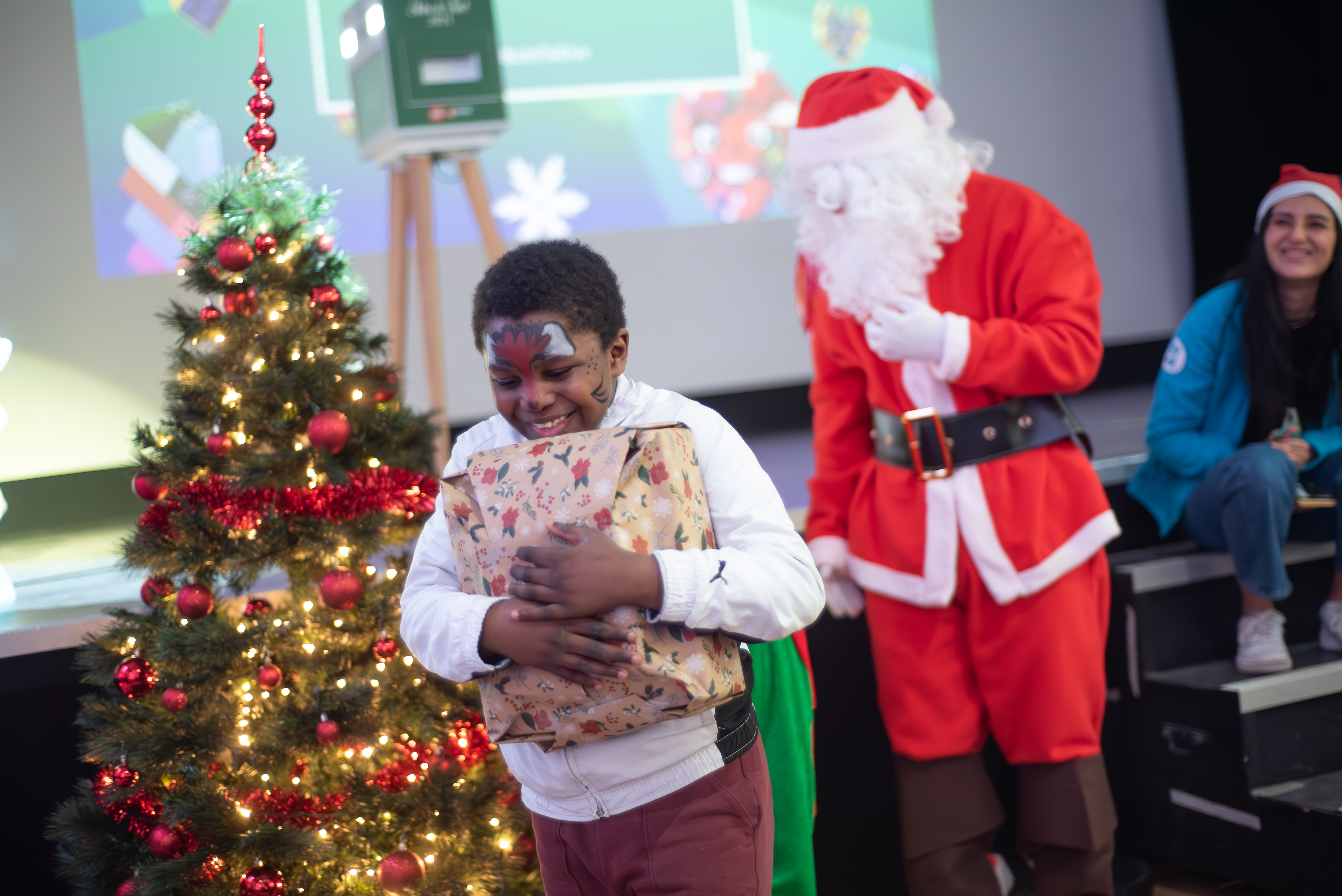 Petit garçon tient un cadeau dans les mains et sourit, derrière un arbre de Noël